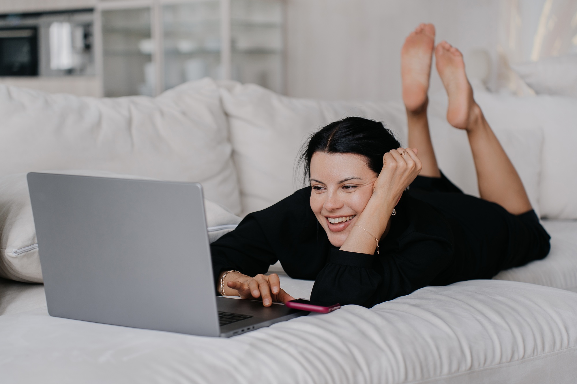 cheerful brunette girl lying on couch home using laptop, smiling, watching comedy movie, lying home